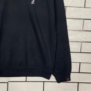 May include: Black sweatshirt with a small white graphic on the upper left chest. The sleeve cuff features a small red tag with a graphic. The sweatshirt is made of a soft material.