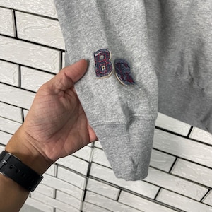 K&ouml;nnte beinhalten: Nahaufnahme eines grauen Sweatshirt-&Auml;rmels mit geripptem B&uuml;ndchen. Der &Auml;rmel hat zwei gestickte Aufn&auml;her mit roten und blauen Akzenten. Die Aufn&auml;her scheinen Buchstaben oder Zahlen zu sein. Der Hintergrund ist eine wei&szlig;e Backsteinmauer.