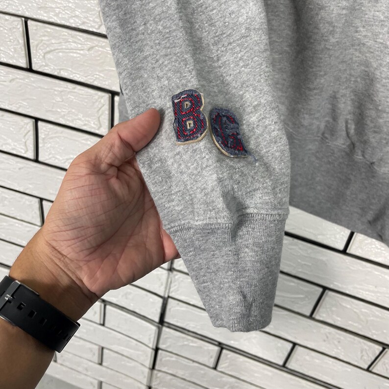 K&ouml;nnte beinhalten: Nahaufnahme eines grauen Sweatshirt-&Auml;rmels mit geripptem B&uuml;ndchen. Der &Auml;rmel hat zwei gestickte Aufn&auml;her mit roten und blauen Akzenten. Die Aufn&auml;her scheinen Buchstaben oder Zahlen zu sein. Der Hintergrund ist eine wei&szlig;e Backsteinmauer.