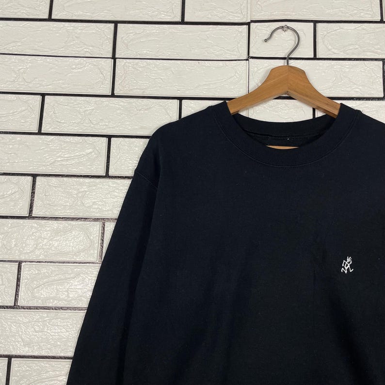 May include: Black crew neck sweatshirt hanging on a wooden hanger. The sweatshirt has a small white embroidered logo on the left chest. The background is a white brick wall.