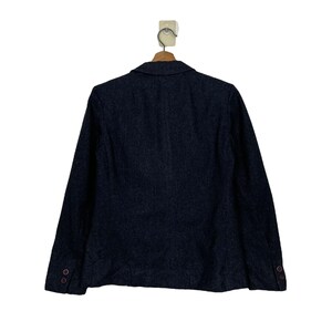 May include: A dark grey blazer with long sleeves, a notched lapel, and two buttons on each cuff. The blazer is hanging on a wooden hanger against a white background. The back of the blazer is visible.