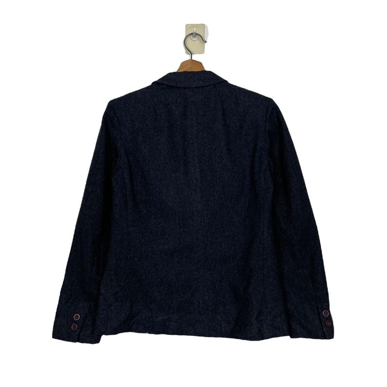 May include: A dark grey blazer with long sleeves, a notched lapel, and two buttons on each cuff. The blazer is hanging on a wooden hanger against a white background. The back of the blazer is visible.
