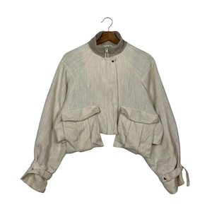 May include: Cream-colored cropped jacket with a high collar and two large front pockets. The jacket has long sleeves with buttoned cuffs and a zipper closure. The fabric has a subtle textured appearance.