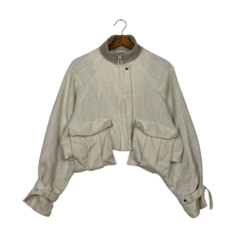 May include: Cream-colored cropped jacket with a high collar and two large front pockets. The jacket has long sleeves with buttoned cuffs and a zipper closure. The fabric has a subtle textured appearance.