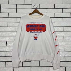 Puede incluir: Sudadera blanca de manga larga con el texto "SERVICE REPAIR / BEST IN TOWN ASSEMBLY SHOP JOHNS Co., Ltd. WORK ORIGINAL GOODS". Tiene un gráfico de dibujos animados y diseños de llaves inglesas rojas en la manga derecha.