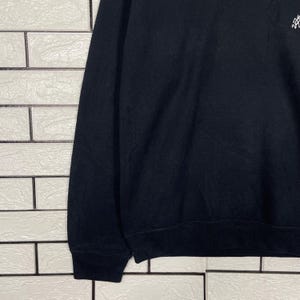 May include: Black sweatshirt with a small, white graphic on the upper right chest. The sweatshirt has long sleeves and a ribbed hem and cuffs. The fabric appears to be a soft, medium-weight material. The background is a white brick wall.