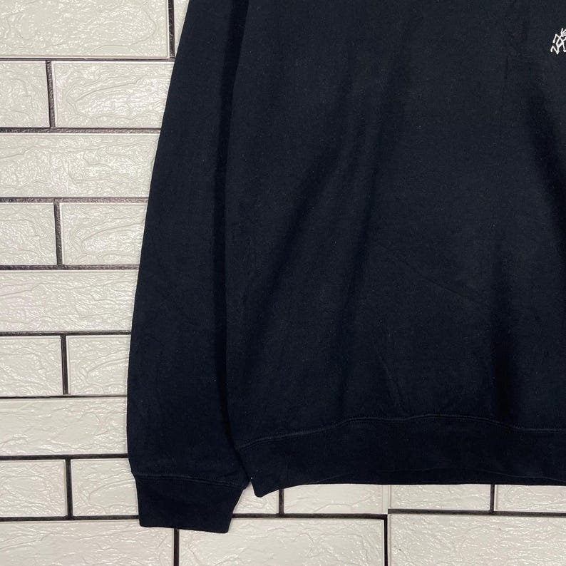 May include: Black sweatshirt with a small, white graphic on the upper right chest. The sweatshirt has long sleeves and a ribbed hem and cuffs. The fabric appears to be a soft, medium-weight material. The background is a white brick wall.