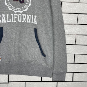 K&ouml;nnte beinhalten: Ein grauer Kapuzenpullover mit dem Wort "CALIFORNIA" in gro&szlig;en wei&szlig;en Buchstaben. Dar&uuml;ber befindet sich ein kreisf&ouml;rmiges Emblem mit der Aufschrift "CALIFORNIA SURF GROOVE". Der Kapuzenpullover hat eine dunkelblaue Tasche und Bes&auml;tze.