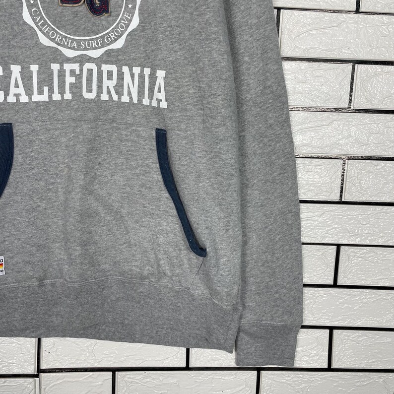 K&ouml;nnte beinhalten: Ein grauer Kapuzenpullover mit dem Wort "CALIFORNIA" in gro&szlig;en wei&szlig;en Buchstaben. Dar&uuml;ber befindet sich ein kreisf&ouml;rmiges Emblem mit der Aufschrift "CALIFORNIA SURF GROOVE". Der Kapuzenpullover hat eine dunkelblaue Tasche und Bes&auml;tze.