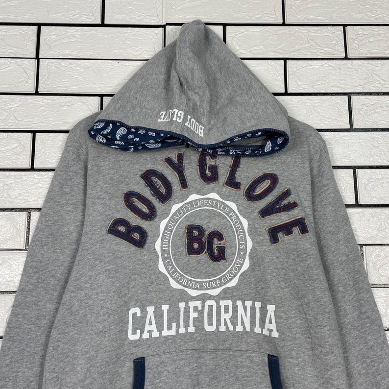 K&ouml;nnte beinhalten: Ein grauer Kapuzenpullover mit dem Text "BODY GLOVE" in dunkelblauer Schrift. Der Pullover hat eine wei&szlig;-blaue Grafik auf der Vorderseite mit den Worten "CALIFORNIA SURF GROOVE". Die Kapuze hat ein blaues Paisley-Muster.