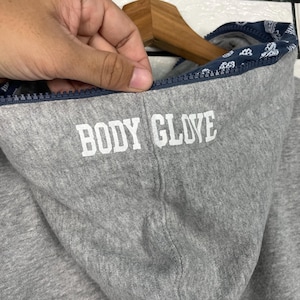 K&ouml;nnte beinhalten: Nahaufnahme eines grauen Kapuzenpullovers mit dem Text "BODY GLOVE" in wei&szlig;en Blockbuchstaben. Die Kapuze hat ein marineblaues Futter mit wei&szlig;em Paisley-Muster. Der Rei&szlig;verschluss ist marineblau.