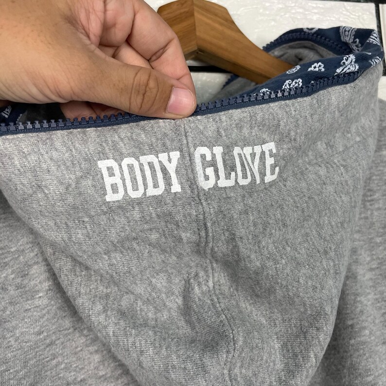 K&ouml;nnte beinhalten: Nahaufnahme eines grauen Kapuzenpullovers mit dem Text "BODY GLOVE" in wei&szlig;en Blockbuchstaben. Die Kapuze hat ein marineblaues Futter mit wei&szlig;em Paisley-Muster. Der Rei&szlig;verschluss ist marineblau.