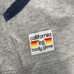 K&ouml;nnte beinhalten: Nahaufnahme eines grauen Sweatshirts mit marineblauer Einfassung. Ein wei&szlig;es rechteckiges Patch zeigt den Text "california bg body glove" mit einem farbenfrohen Sonnenuntergangsdesign.
