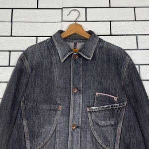 May include: A dark grey denim jacket with a button-up front, a collared neckline, and a single chest pocket. The jacket is hanging on a wooden hanger against a white brick wall.