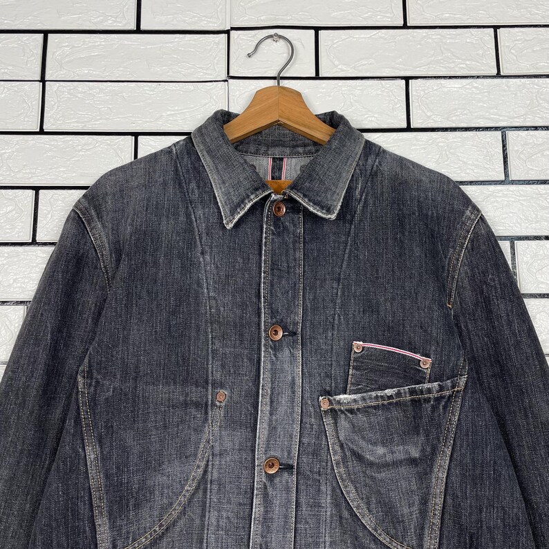 May include: A dark grey denim jacket with a button-up front, a collared neckline, and a single chest pocket. The jacket is hanging on a wooden hanger against a white brick wall.