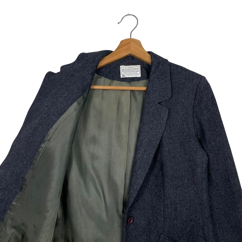 May include: A dark grey blazer with a single-button fastening, suspended from a wooden hanger. The interior lining is a muted olive green. A label is visible near the collar. The blazer is made of a textured fabric.