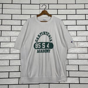 Puede incluir: Sudadera blanca de manga corta con el texto "CARPINTERIA 85 S 4 ACADEMY" en verde. La sudadera tiene un pequeño logotipo de Champion en la manga izquierda. La prenda está colgada en una percha de madera.