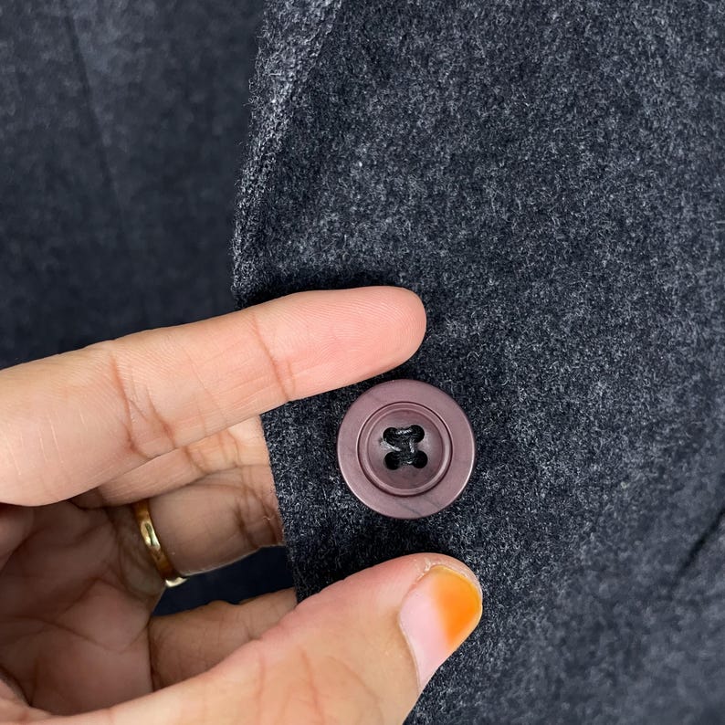 May include: Close-up of a dark grey wool blazer with a round, mauve button. The button is centered on the blazer's fabric, and the texture of the wool is visible. The button has a four-hole design.