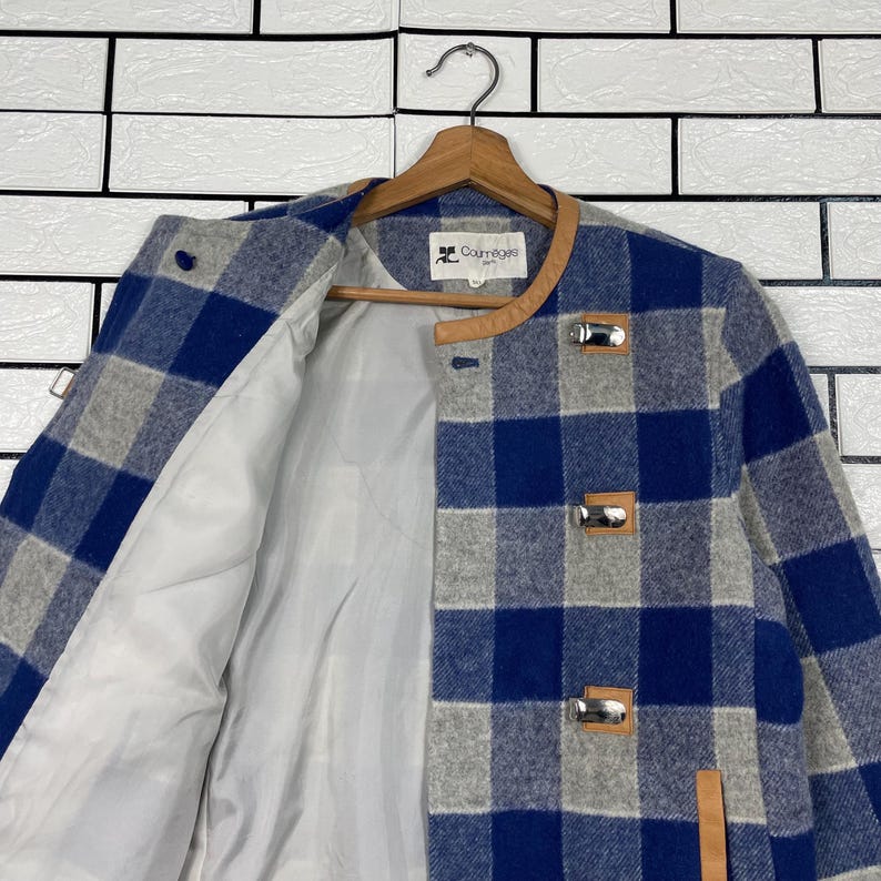 May include: A blue and grey checked jacket with a light lining. The jacket features a round collar with brown trim and silver clasps. It is suspended on a wooden hanger.