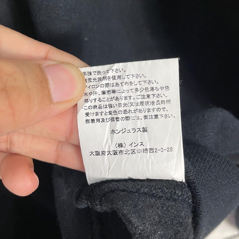 May include: A close-up of a black garment tag with Japanese text. The tag is white with black text, and the garment is black. The tag provides washing and care instructions.