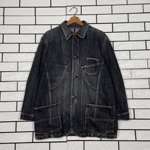 May include: A dark grey denim jacket with a button-up front, two large front pockets, and a small chest pocket. The jacket is hanging on a wooden hanger against a white brick wall.