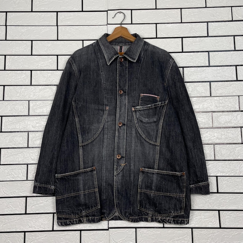 May include: A dark grey denim jacket with a button-up front, two large front pockets, and a small chest pocket. The jacket is hanging on a wooden hanger against a white brick wall.