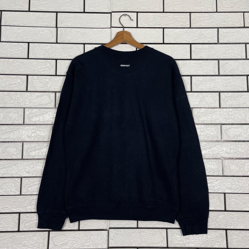 May include: A black long-sleeve sweatshirt with a small white logo near the neckline. The sweatshirt is hanging on a wooden hanger against a white brick wall. The logo appears to read "409-427".