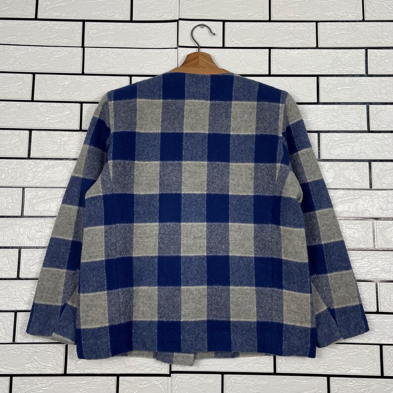 May include: A blue, grey, and brown plaid jacket hanging on a wooden hanger. The jacket features a classic plaid pattern in shades of blue and grey. The collar is brown. The jacket is displayed against a white brick wall.