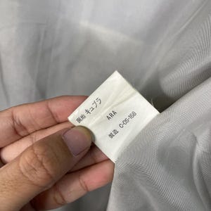 May include: A close-up of a white tag with Japanese text and the word "ARA" printed in black. The tag also has the text "C-OS-168". The tag is held by a person's hand and is against a grey fabric.