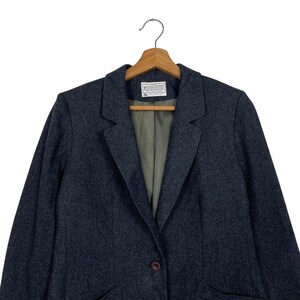 May include: A dark grey blazer with a single button fastening, suspended on a wooden hanger. The blazer features a traditional lapel design and an internal label. The material seems to be a wool blend.