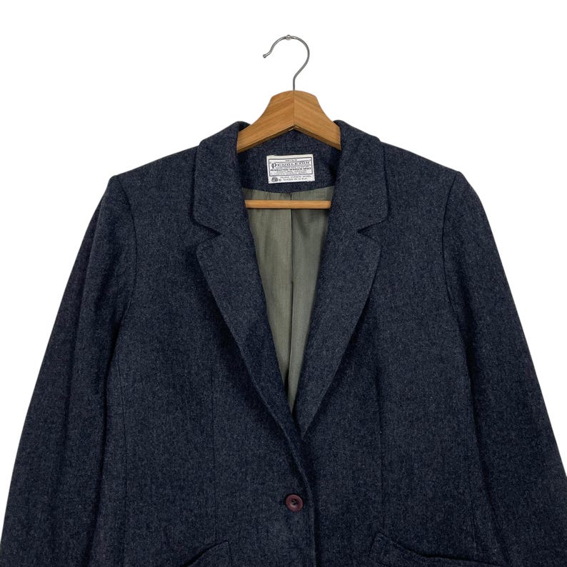 May include: A dark grey blazer with a single button fastening, suspended on a wooden hanger. The blazer features a traditional lapel design and an internal label. The material seems to be a wool blend.