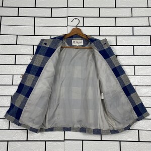 May include: A blue, grey, and white checked jacket with a light grey interior lining. The jacket is suspended on a wooden hanger. The jacket has a button closure and toggle clasps. The background is a white brick wall.