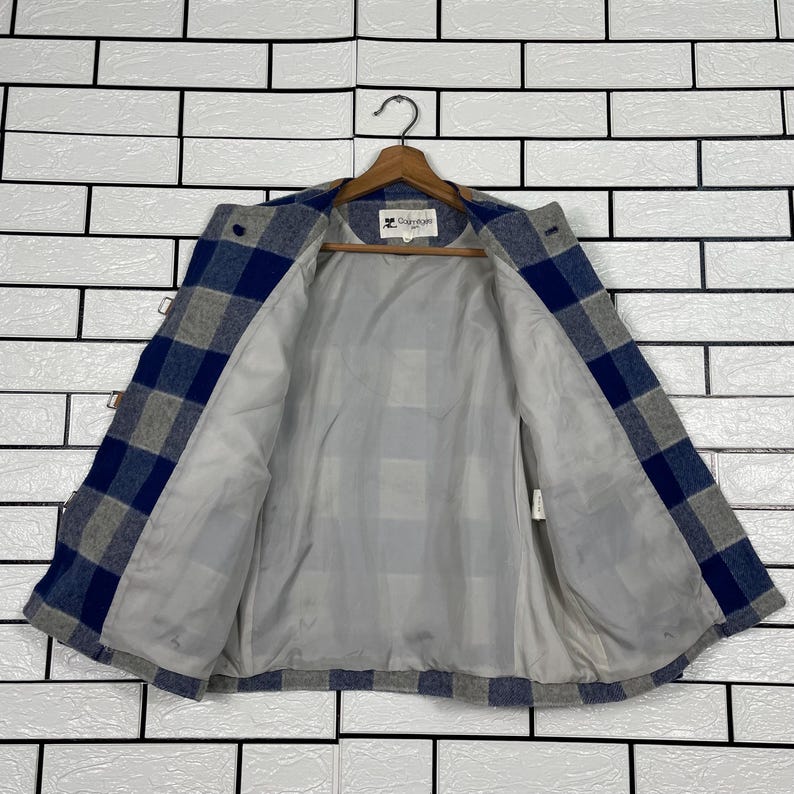 May include: A blue, grey, and white checked jacket with a light grey interior lining. The jacket is suspended on a wooden hanger. The jacket has a button closure and toggle clasps. The background is a white brick wall.