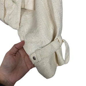 May include: Close-up of a cream-colored woven fabric sleeve. The sleeve features a buttoned cuff with a decorative loop detail. The fabric has a textured appearance, and the sleeve is slightly gathered at the shoulder.