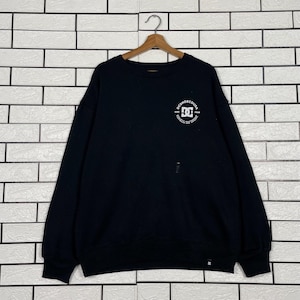 May include: Black crew neck sweatshirt with the DC Shoes logo in white on the upper left chest. The sweatshirt is hanging on a wooden hanger against a white brick wall. The DC Shoes logo is a stylized design.