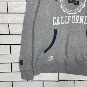 K&ouml;nnte beinhalten: Grauer Kapuzenpullover mit dem Wort "CALIFORNIA" in gro&szlig;en wei&szlig;en Buchstaben. Der Pullover hat eine K&auml;ngurutasche und marineblaue Akzente an der Kapuze. Das Markenlogo ist oben sichtbar.