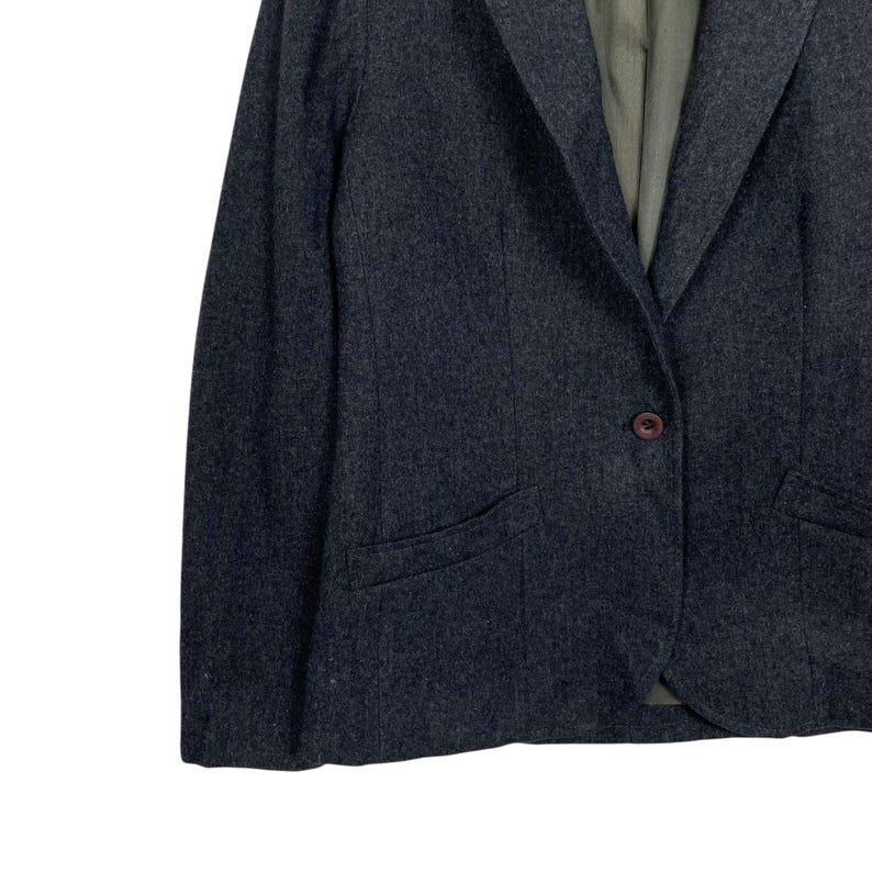 May include: A dark grey blazer with a single-button fastening and a notched lapel. The blazer features two front pockets and a subtle textured pattern. The interior lining is a muted green colour. The overall style is classic and tailored.