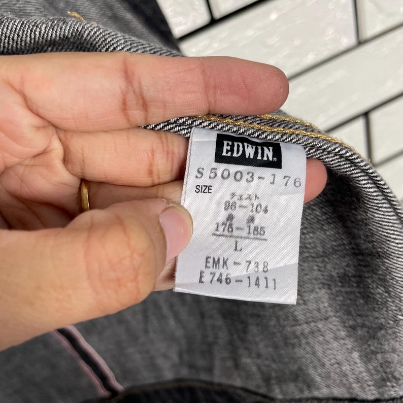 May include: A close-up of a grey denim jacket with an Edwin brand label. The label shows the model number S5003-176, size L, and Japanese text. The jacket's fabric is visible, showing a textured weave.
