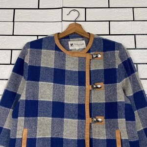 May include: A blue, grey, and white checked jacket with brown leather trim and silver clasps. The jacket is suspended on a wooden hanger. The brand name "Courr&egrave;ges" is visible on the collar. The background is a white brick wall.