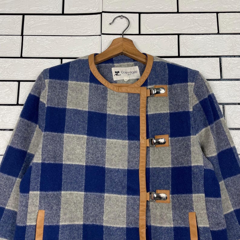 May include: A blue, grey, and white checked jacket with brown leather trim and silver clasps. The jacket is suspended on a wooden hanger. The brand name "Courr&egrave;ges" is visible on the collar. The background is a white brick wall.