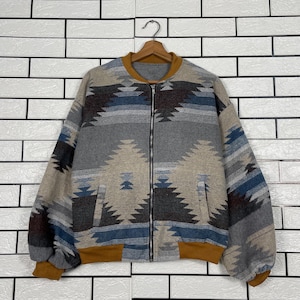 May include: A zip-up jacket with a geometric pattern in shades of gray, blue, and tan. The jacket has a brown collar, cuffs, and hem. The jacket is hanging on a wooden hanger against a white brick wall.