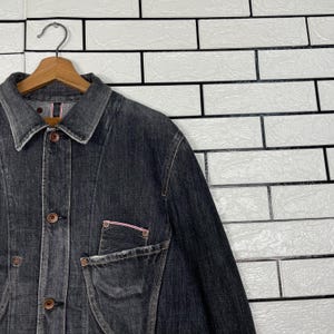 May include: A dark grey denim jacket with a button-up front and a collared neckline. The jacket has a chest pocket with a pink trim and is hanging on a wooden hanger. The background is a white brick wall.