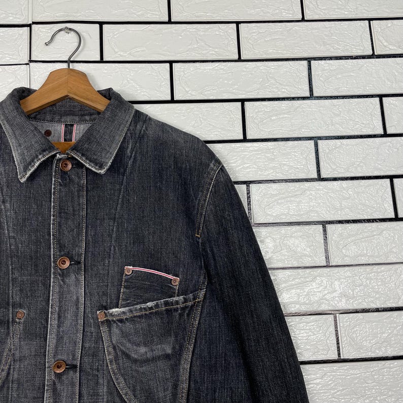May include: A dark grey denim jacket with a button-up front and a collared neckline. The jacket has a chest pocket with a pink trim and is hanging on a wooden hanger. The background is a white brick wall.
