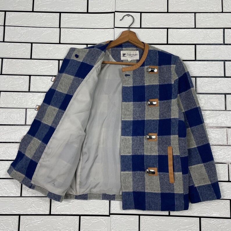 May include: A blue, grey, and white checked jacket with a light grey lining. The jacket features a brown leather collar and toggle fastenings. It is displayed on a wooden hanger.