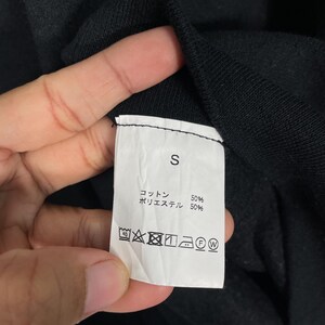 May include: Close-up of a white clothing tag with the letter "S" printed in black. The tag also includes Japanese text and washing instructions. The fabric composition is 50% cotton and 50% polyester.