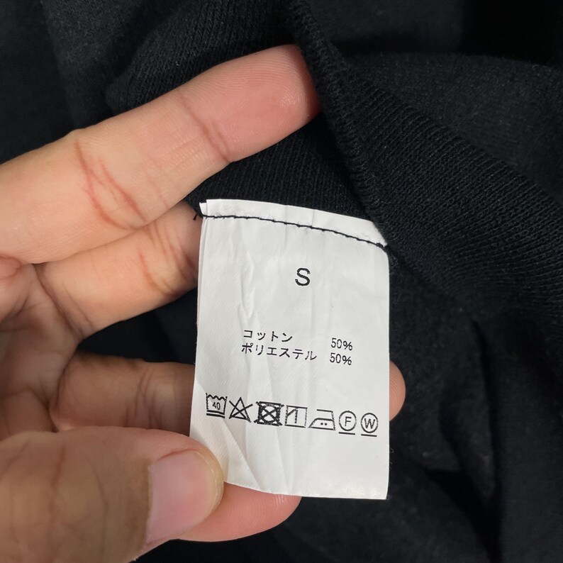 May include: Close-up of a white clothing tag with the letter "S" printed in black. The tag also includes Japanese text and washing instructions. The fabric composition is 50% cotton and 50% polyester.