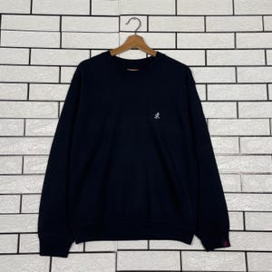 May include: A navy blue sweatshirt with a small white logo on the chest. The long-sleeved top is hanging on a wooden hanger against a white brick wall.