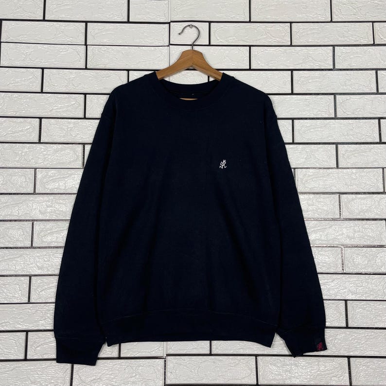 May include: A navy blue sweatshirt with a small white logo on the chest. The long-sleeved top is hanging on a wooden hanger against a white brick wall.