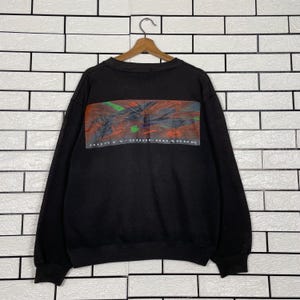 May include: Black sweatshirt with a rectangular graphic on the back featuring a colorful abstract design in red, green, and black. The text "RUSTY-SURF BOARDS" is printed below the graphic. The sweatshirt is hanging on a wooden hanger.