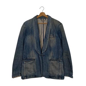 Peut inclure: Un blazer en jean bleu avec un col châle, deux poches avant et une fermeture à un bouton. La veste est suspendue à un cintre en bois sur fond blanc. Le blazer a un délavage décoloré et une coupe décontractée.